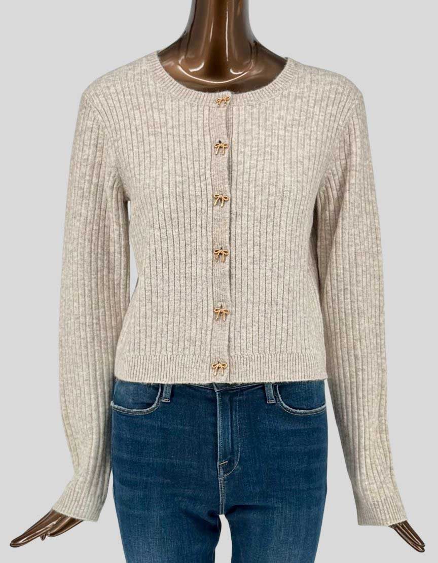  Authentic screen-worn Icône par Simons cream ribbed-knit cardigan featuring charming gold-tone bow button details. Classic crew neck design with long sleeves and fitted silhouette. 