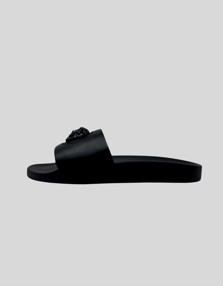 VERSACE Palazzo Medusa Pool Slide Sandals in Black. Iconic three-dimensional Medusa plaque at center. Surrounded by signature Greca pattern accent. All-black rubber construction. Molded footbed with Medusa head logo. Slip-on style.