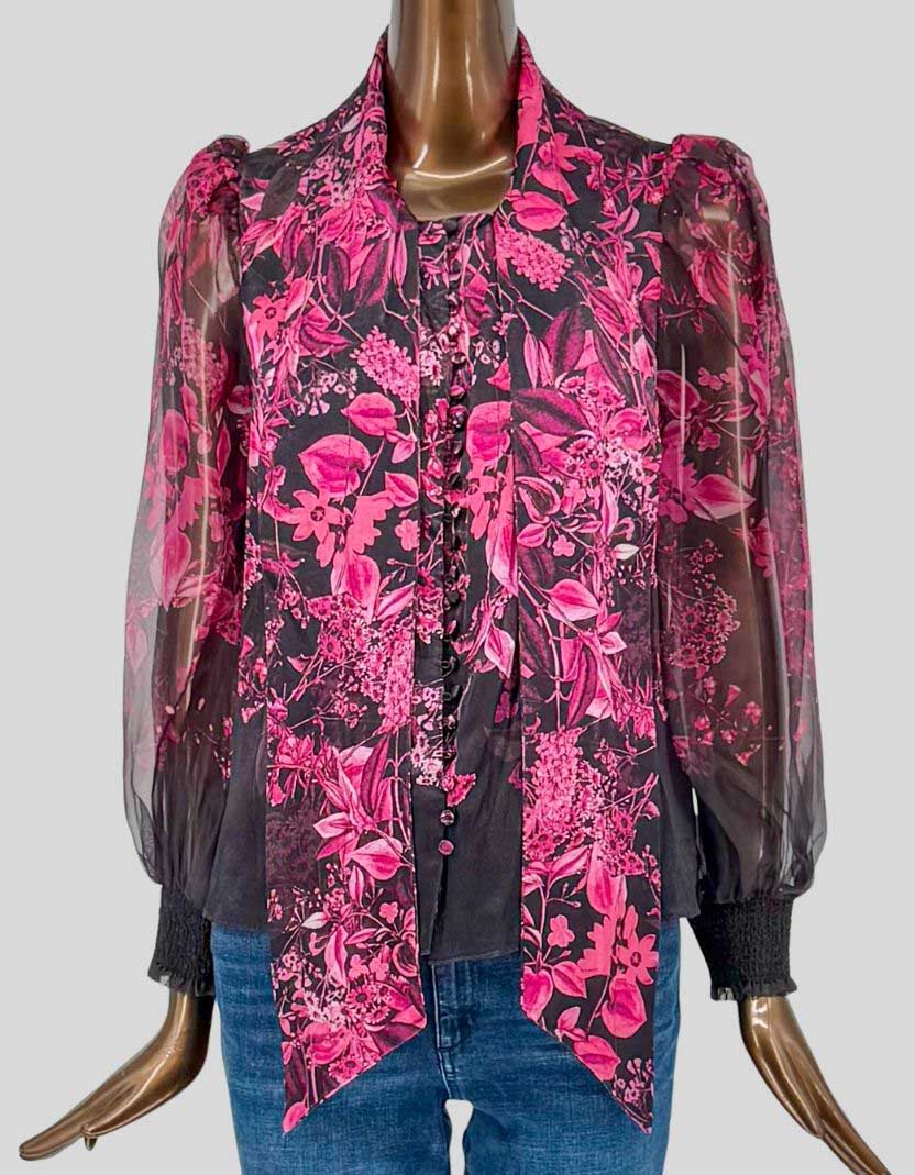 ALICE + OLIVIA dramatic floral blouse in vibrant pink botanical print on black background. Sheer silk-blend with integrated tie-neck bow collar. Long blouson sleeves with smocked elastic cuffs.
