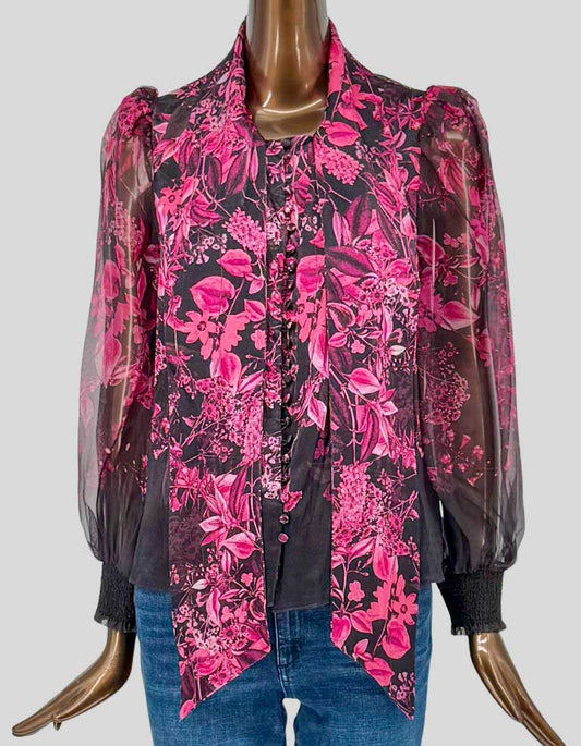 ALICE + OLIVIA dramatic floral blouse in vibrant pink botanical print on black background. Sheer silk-blend with integrated tie-neck bow collar. Long blouson sleeves with smocked elastic cuffs.