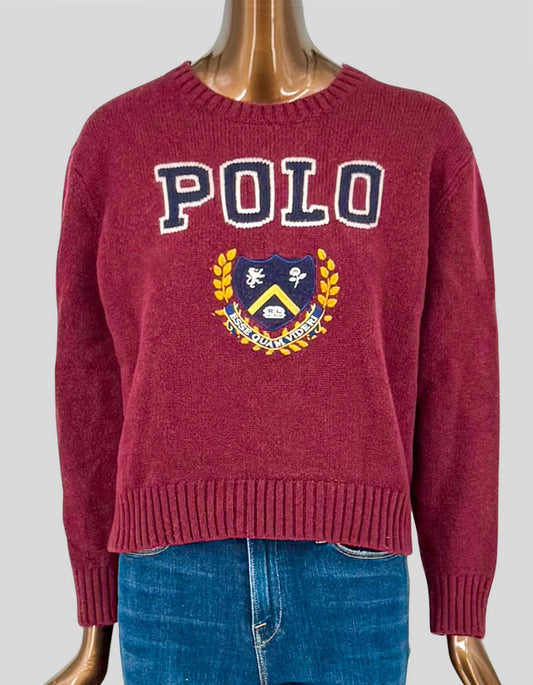 POLO RALPH LAUREN crew neck sweater in rich burgundy featuring bold "POLO" lettering and classic crest emblem at front. Made from 100% wool for warmth and durability. The collegiate-inspired graphic design showcases iconic Polo Ralph Lauren branding with embroidered crest details. Ribbed crew neck, cuffs, and hem provide structure and classic finishing. A preppy classic.